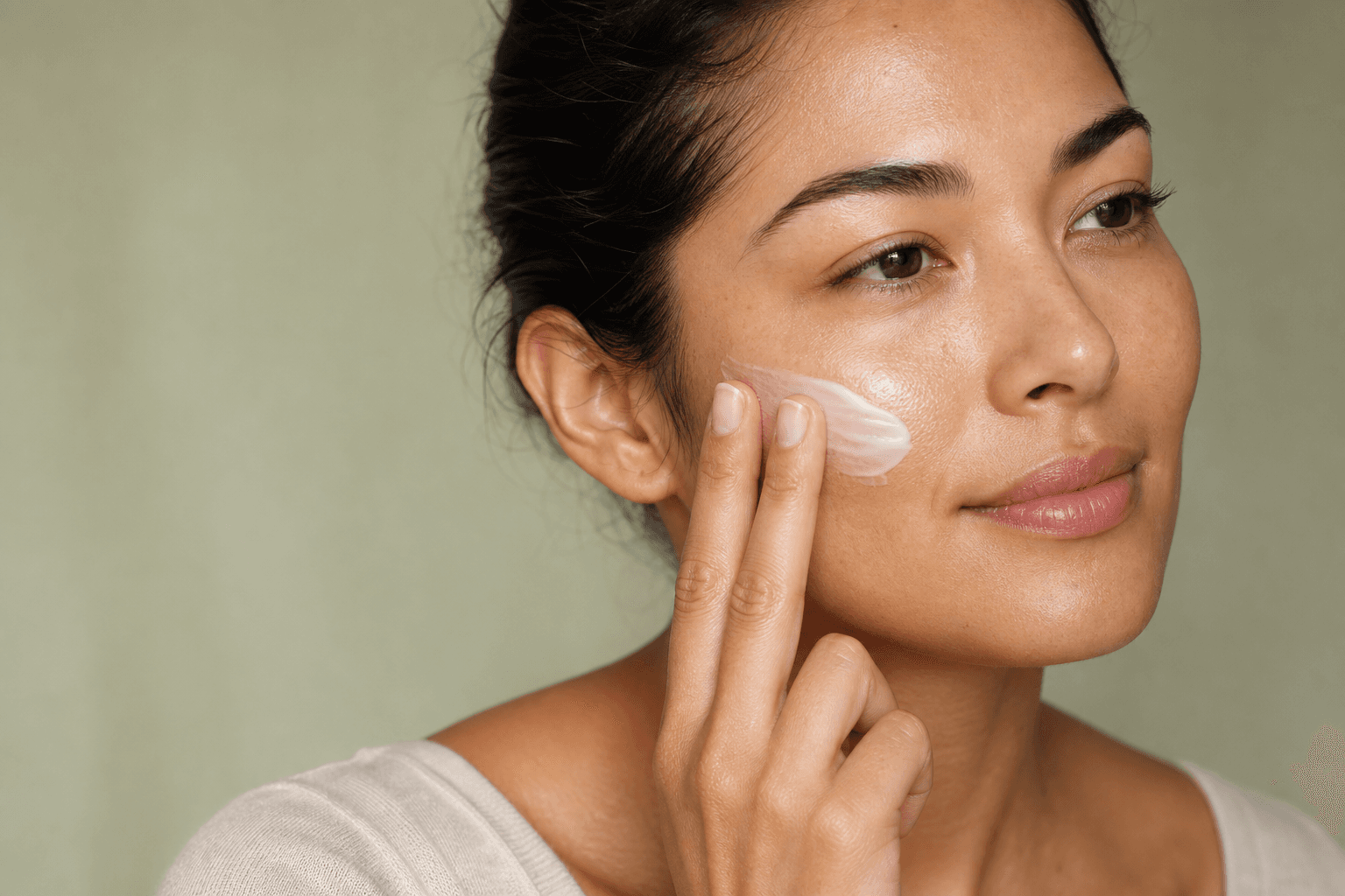 Woman with natural skin texture applying white moisturizer to her cheek